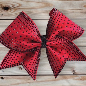 May include: A large red cheer bow with black rhinestones. The bow is made of satin and has a pointed shape.