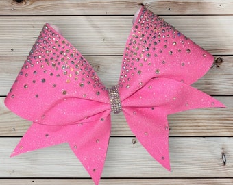 May include: A pink cheer bow with a sparkly finish and a silver rhinestone design. The bow is tied in a classic cheer bow shape.