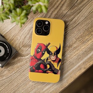 May include: Yellow phone case featuring Deadpool and Wolverine characters making a heart shape with their hands. The case has a glossy finish and a camera cutout. The background is a wooden surface.