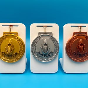Könnte beinhalten: Drei runde Medaillen in Gold, Silber und Bronze, die jeweils auf einem weißen Ständer präsentiert werden. Die Medaillen zeigen ein zentrales Maiskolben-Motiv, umgeben von einem Lorbeerkranz. Die Goldmedaille trägt die Aufschrift CHAMPION, Silber 2ND PLACE und Bronze 3RD PLACE.