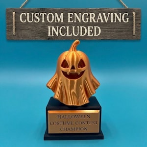 May include: A metallic orange 3D-printed ghost trophy with a carved jack-o'-lantern face sits on a black base. The base features a gold plaque reading HALLOWEEN COSTUME CONTEST CHAMPION. Above, a wooden sign reads CUSTOM ENGRAVING INCLUDED.