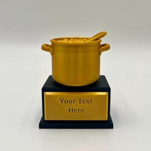 May include: A gold-coloured trophy featuring a pot of food with a spoon, resting on a black base with a gold plaque. The plaque reads "Your Text Here". The pot has two handles.