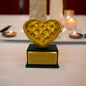 May include: A heart-shaped, gold-colored award or trophy with a woven design on top, mounted on a black base with a gold plate that reads "Your Text Here". The background includes lit candles.