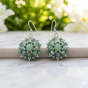 May include: A pair of beaded earrings with a floral design. The earrings feature a central light green stone surrounded by smaller, iridescent green beads. The earrings hang from silver-colored hooks, with a blurred green and white background.