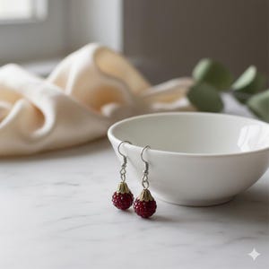 May include: A pair of dangle earrings with red, jewel-like spheres. The earrings have silver-colored hooks and gold-colored caps. The earrings are displayed next to a white bowl on a marble surface.