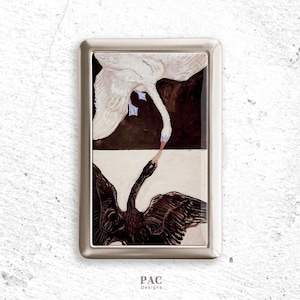 May include: A rectangular, silver-toned metal case featuring a painting of two swans. One swan is white, the other black, with their beaks nearly touching. The artwork is set against a dark brown and cream background. The text "PAC Designs" is at the bottom.