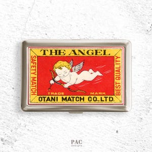 May include: A silver metal match case with a vintage design. The red background features a cherub with a bow and arrow, and the text "THE ANGEL". The case also has the text "SAFETY MATCH" and "BEST QUALITY".