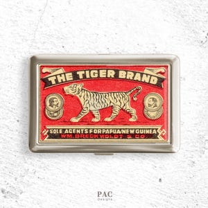 Może przedstawiać: Prostokątne metalowe etui na papierosy z czerwono-czarnym wzorem. Na etui znajduje się napis "THE TIGER BRAND" nad ilustracją tygrysa. Na dole etui widnieje napis "SOLE AGENTS FOR PAPUA/NEW GUINEA".