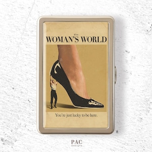 May include: A rectangular metal cigarette case featuring a vintage-style graphic of a person holding up a large black high-heeled shoe. The text reads It's a WOMAN'S WORLD and You're just lucky to be here. The case has a silver-toned metallic frame.