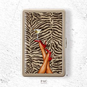 Peut inclure: Un étui à cigarettes rectangulaire en métal avec une impression graphique représentant des jambes portant des chaussures à talons hauts rouges, équilibrant un verre à martini sur les pieds, sur un motif zébré marron et noir avec le texte PAC Designs.
