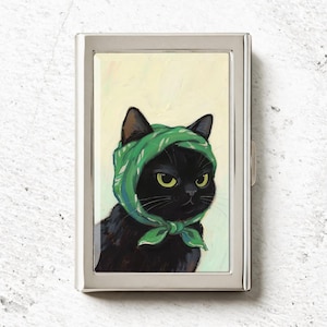May include: A rectangular silver metal case featuring an illustration of a black cat wearing a green headscarf tied under its chin. The cat has yellow-green eyes and is set against a pale yellow background. The case has a polished metallic frame.