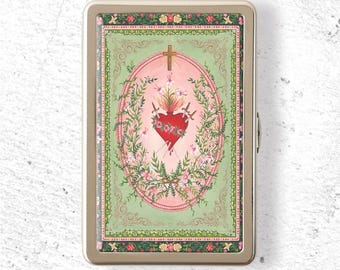 May include: A rectangular metal cigarette case featuring a gold-toned frame and a central illustration of a red heart pierced by two swords, surrounded by a floral wreath and a cross, set against a pale pink oval and a light green background.