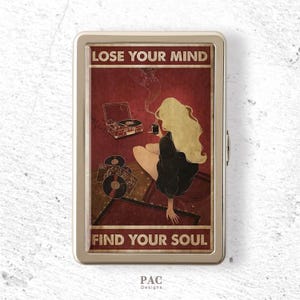 Op de afbeelding: Een rechthoekig metalen sigarettendoosje met een vintage-stijl illustratie van een persoon met lang blond haar zittend bij een platenspeler en vinylplaten. De tekst LOSE YOUR MIND en FIND YOUR SOUL staat in het wit op een donkerrode achtergrond.
