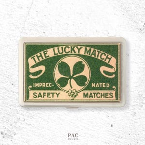 May include: A rectangular metal case featuring a green and cream label with the text THE LUCKY MATCH, IMPREG-NATED, and SAFETY MATCHES. The design includes a central four-leaf clover graphic inside a circle, framed by a silver-toned metal border.