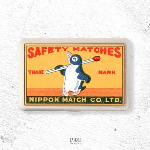 May include: A rectangular metal case featuring a vintage-style label for Safety Matches. The label has a yellow background with red text, a graphic of a penguin carrying a match, and the text Nippon Match Co., Ltd. printed in black.