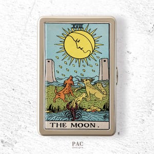 May include: A rectangular metal case featuring a graphic print of The Moon tarot card. The design displays a yellow sun, two towers, a dog, a wolf, and a lobster in a blue and green landscape. The text XVIII and THE MOON are clearly visible.