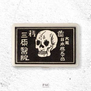 May include: A rectangular metal case with a black and white graphic featuring a central human skull illustration. The design includes Japanese text characters arranged vertically on both sides of the skull, framed by a thin silver-toned border.