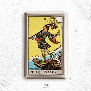 以下が含まれることがあります： 「愚者」のタロットカードイラストが描かれたシルバーのメタルケース。カードには、杖を持った人物、小さな犬、太陽が黄色の背景に描かれています。「THE FOOL」の文字が下部にあります。