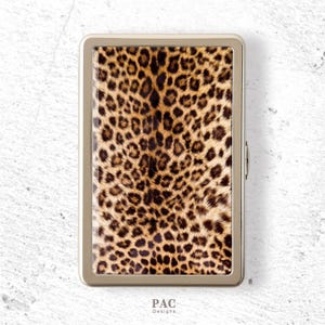 May include: A rectangular metal cigarette case featuring a prominent leopard print pattern with black spots on a tan and brown background. The case has a polished gold-tone metal frame and a side hinge mechanism. The text PAC Designs is printed below.