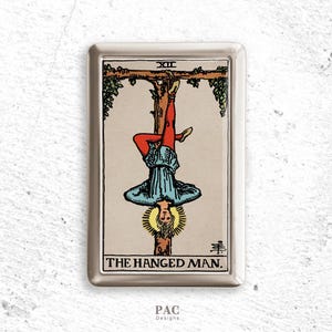 Könnte beinhalten: Ein silbernes Metallgehäuse mit einer Tarotkartenillustration des Gehängten. Die Karte zeigt eine Figur, die kopfüber hängt, mit roter Hose und blauem Hemd, mit einem gelben Heiligenschein. Der Text "THE HANGED MAN" steht unten.