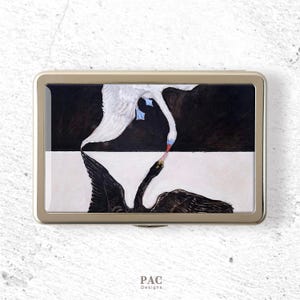 May include: A rectangular metal case with a gold-toned frame featuring a printed design of a white swan and a black swan facing each other. The background is split horizontally into black and white sections. The text PAC Designs is printed below.