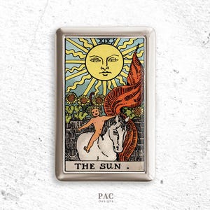 May include: A silver metal case featuring a tarot card design. The card depicts a radiant sun with a smiling face, a child riding a white horse, and the text "THE SUN." The design includes sunflowers and a red banner.