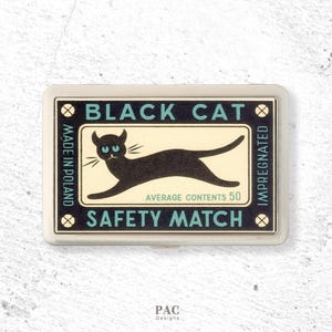 May include: A rectangular metal matchbox case featuring a black cat illustration on a cream background. The text reads BLACK CAT, SAFETY MATCH, MADE IN POLAND, IMPREGNATED, and AVERAGE CONTENTS 50 in teal and black lettering with small gold accents.