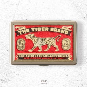 Peut inclure: Un étui métallique rectangulaire avec une étiquette rouge illustrée d'un tigre noir. Le texte indique THE TIGER BRAND, SOLE AGENTS FOR PAPUA NEW GUINEA et WM. BRECKWOLDT & CO. Deux médaillons circulaires avec des portraits flanquent le tigre.