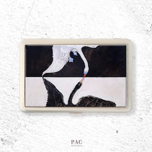 May include: A rectangular artwork depicting two swans, one black and one white, with their beaks touching. The background is split, with a black upper half and a white lower half. The piece is framed in a cream-colored border.
