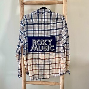 Könnte beinhalten: Ein weißes, blaues und orangefarbenes kariertes Flanellhemd mit einem schwarzen Patch auf der Rückseite, auf dem "ROXY MUSIC" in weißen Buchstaben steht.