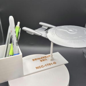 Pode incluir: Um organizador de mesa branco com um porta-canetas e um pequeno modelo da nave espacial USS Enterprise de Star Trek. A base do modelo da nave espacial tem o texto "NCC-1701-D USS ENTERPRISE".