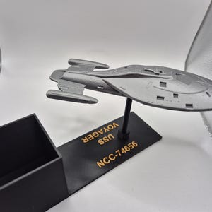 Könnte beinhalten: Ein graues Modell des Raumschiffs USS Voyager aus Star Trek. Das Modell steht auf einem schwarzen Sockel mit dem aufgedruckten Text "NCC-74656 USS VOYAGER".