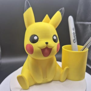 May include: A yellow Pikachu-shaped desk organizer with a matching yellow pen holder. The Pikachu figure has black eyes, red cheeks, and a smiling red mouth. A white Sharpie pen is in the pen holder.