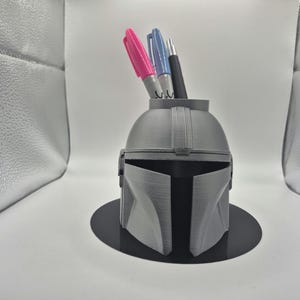 May include: A gray plastic Mandalorian helmet shaped desk organizer with three pens inside.