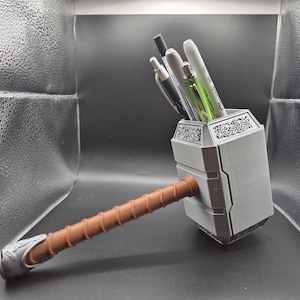May include: A grey and brown Mjolnir-shaped desk organiser with a decorative design. The organiser is filled with pens and pencils.