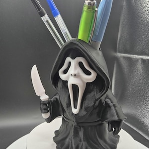 Puede incluir: Un portalápices en blanco y negro con la forma del personaje Ghostface de la serie de películas de terror Scream. El portalápices sostiene varios bolígrafos y un cuchillo blanco. El portalápices está sobre una superficie blanca.