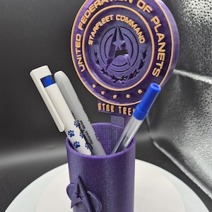 May include: A purple pen holder with a Star Trek theme. The holder features the Starfleet Command emblem and the words "United Federation of Planets." It holds a white marker, a gray marker, and a blue pen. The holder is on a white surface.