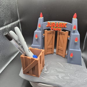 May include: A gray and brown 3D printed Jurassic Park gate with a wooden crate pencil holder. The gate has a sign that reads "Jurassic Park" and has red flames on the top of the towers.