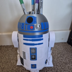 Op de afbeelding: Een witte, blauwe en grijze pennenhouder in de vorm van het Star Wars-personage R2-D2. De pennenhouder heeft een cilindrisch lichaam met blauwe en grijze details en bevat verschillende Sharpie-pennen in diverse kleuren.