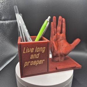 May include: A red desk tidy with the phrase "Live long and prosper" on the front. The organiser holds pens and features a hand gesture. The organiser is made of a reddish-brown material.