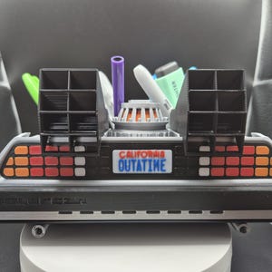 May include: A detailed model of a DeLorean time machine, featuring a black and silver body with orange and red tail lights. The license plate reads "CALIFORNIA OUTATIME". The model includes a central reactor and two black vents.