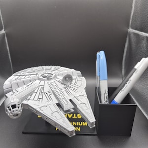 Op de afbeelding: Een gedetailleerd, grijs Millennium Falcon-model staat op een zwarte basis met tekst. Een zwarte pennenhouder bevat een blauwe en een grijze Sharpie-marker. De scène speelt zich af tegen een donkere achtergrond.