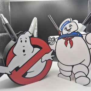 Może przedstawiać: Organizery na biurko z postaciami z Ghostbusters. Jednym z nich jest logo Ghostbusters z duchem, drugim - Stay Puft Marshmallow Man. Długopisy są widoczne w organizerach. Organizery są białe, czerwone i czarne.