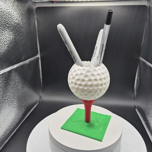 May include: A white golf ball-shaped pen holder with a dimpled surface, resting on a red tee and a green square base. Two pens and a marker are inside the holder. The golf-themed desk accessory is on a white platform.