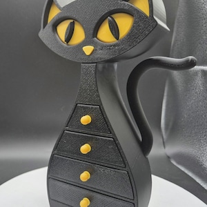 Könnte beinhalten: Eine schwarze, katzenförmige Schmuckschatulle mit gelben Augen, Ohren und Schubladengriffen. Die Katze hat eine strukturierte Oberfläche und einen gebogenen Schwanz. Die Box hat vier Schubladen.