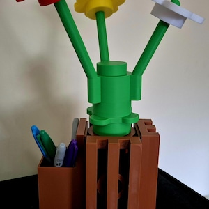 May include: A 3D-printed desk organizer featuring a flower design. The organizer has three colorful flowers: red, yellow, and white, with green stems. A brown pen holder is attached to the base, holding several pens.