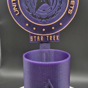 May include: A purple Star Trek themed desk organiser. The top features the Starfleet Command emblem and text. The base is a cylindrical holder with the Starfleet emblem. The text "STAR TREK" is visible.