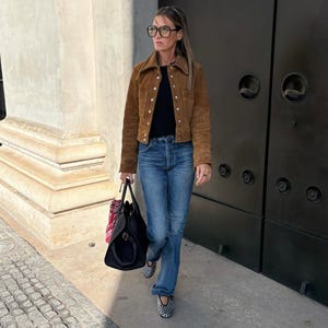 May include: A woman wearing a brown suede jacket, black top, and blue jeans. She carries a black handbag and wears studded flats. The outfit is completed with eyeglasses and a headband. The background includes a stone wall and a dark door.