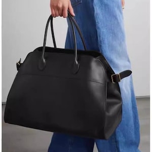 Peut inclure: Un grand sac fourre-tout en cuir noir avec deux poignées supérieures et une bandoulière. Le sac a une forme rectangulaire avec des bords arrondis et une fermeture à boucle. Le sac est porté par une personne portant un jean bleu.