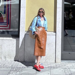 May include: A woman wearing a light blue button-down shirt, a white top, and a brown leather skirt. She is also wearing red shoes and sunglasses, and carrying a straw bag. The outfit is set against a yellow wall and a stone walkway.